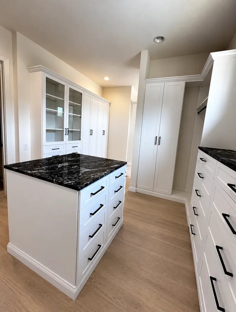 White walk-in closet with glass-front upper cabinets, drawer banks and a black granite island