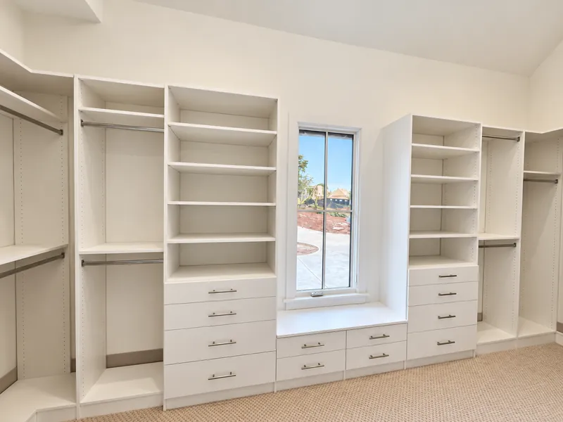 White walk-in closet with shelving and hanging rods on both sides of a window, with drawers beneath it