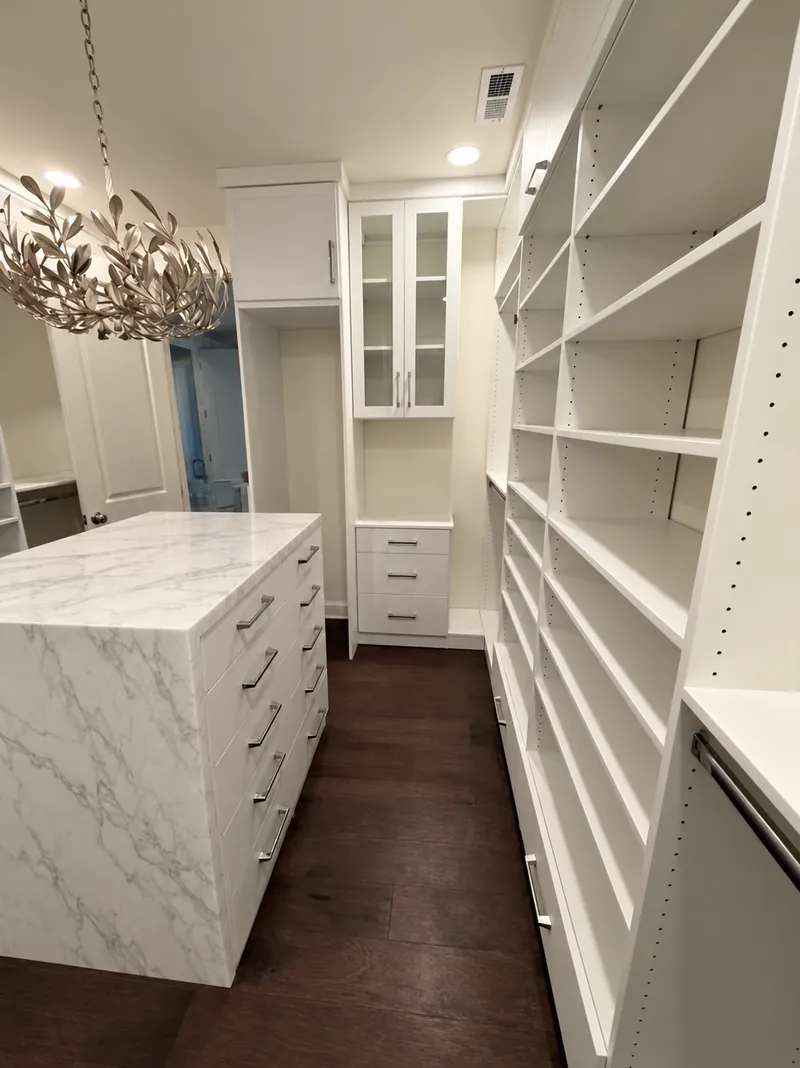 Walk-in closet aisle with floor-to-ceiling white shelving, a glass-front cabinet and a marble island